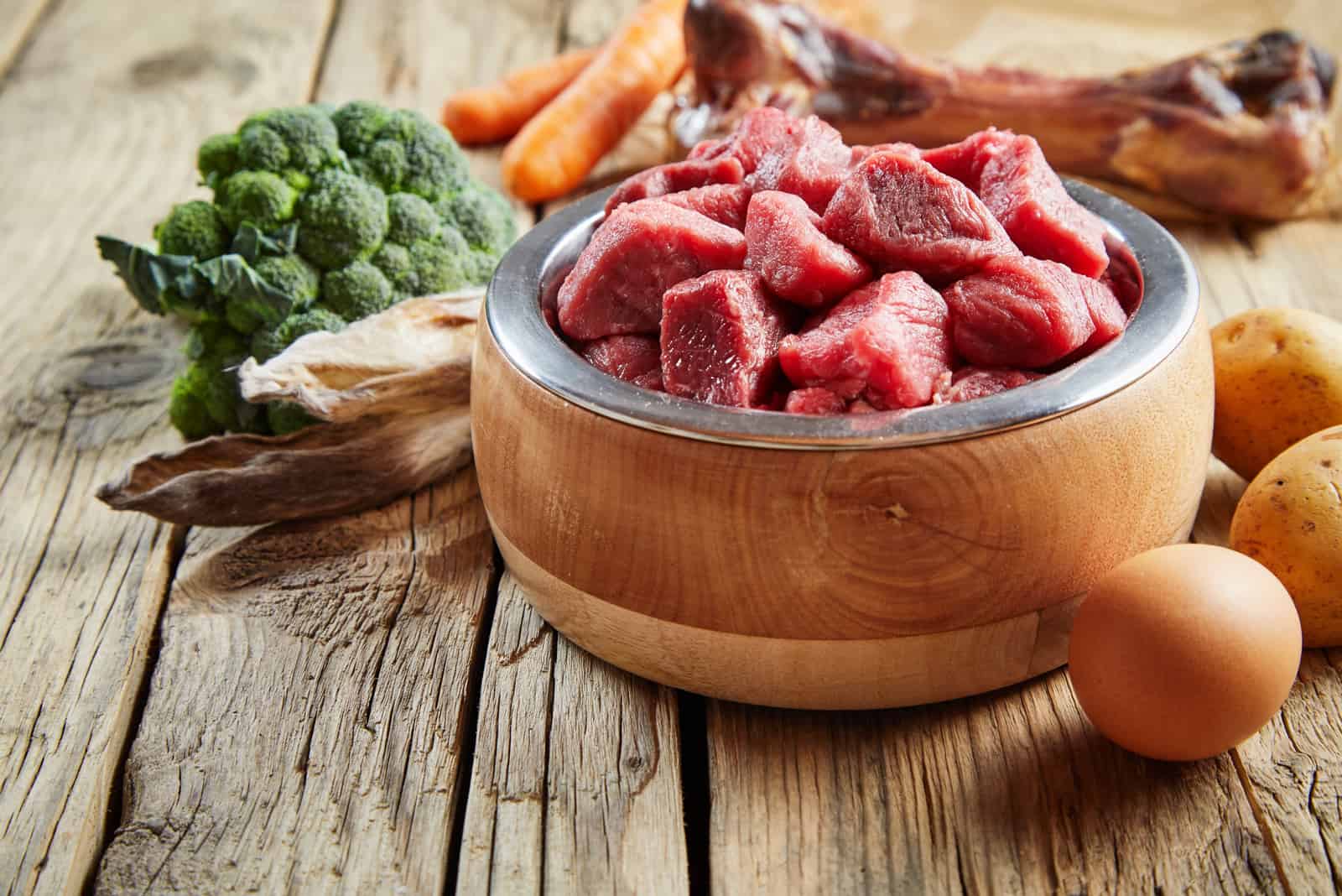 Frisches Futter für ein gesundes Haustier mit gehacktem Rindfleisch, Eiern, Brokkoli, Gemüse und einem großen Fleischknochen auf einem rustikalen Holzboden